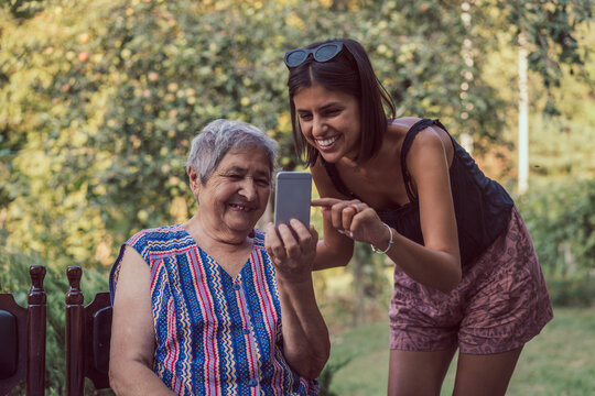 Young Woman And Very Old Happy Woman Using Smartphone Outdoors. New Generations Teaching Old Generations To Use New Technologies Mobile Phone