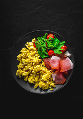 scrambled eggs with prosciutto and salad on black plate on Dark grey black slate background