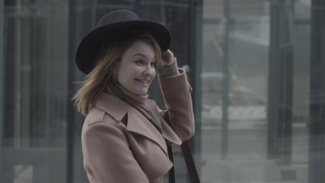 Side view of an elegant happy woman in coat and hat walking in the street. Action. Laughing woman taking off a hat because of strong wind.