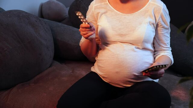 Pregnant woman holding blister packs with drugs sits on a sofa in living room and sighs in close-up. Young lady hesitates and stresses if meds can hurt her baby. Healthcare and future motherhood theme