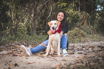 Mujer sentada en la tierra con perro labrador color crema. Labrador con lazo rojo. Mujer con Lentes y Camisa negra con roja.