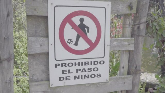 A door with a forbidden white sign and symbol in Spanish that translates "Children passing forbidden" prevents kids entering a small lake in a park.
