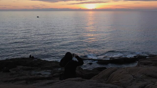 Girl taking a Picture of the Sea and the Sunset with her Smartphone and Lovers Sitting on the Rocks in front of the Sea