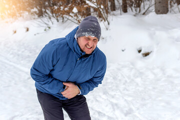 Fototapeta na wymiar Young handsome caucasian man wearing cap and jacket on snowy weather with hand on stomach causing pain in stomach, feeling unwell. Photo of young man with stomach pain. Frustrated man with stomachache