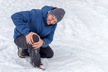 Photo of injured young man bend down on snow and holding his knee in pain, outdoor. Young man holding his knee in pain on a snowy cold winter day. Man having a knee injury on winter road. Copy space.