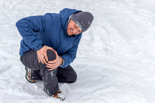 Photo of injured young man bend down on snow and holding his knee in pain, outdoor. Young man holding his knee in pain on a snowy cold winter day. Man having a knee injury on winter road. Copy space.