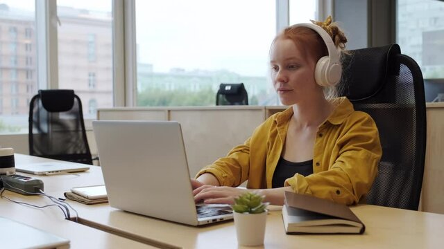 Medium shot of young woman working on laptop in contemporary office then putting on headphones and continuing working
