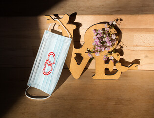 Valentine's Day celebration concept. medical protective mask with drawn hearts, wooden inscription the word love on a sunlit beige background. Positive thinking in covid 19 pandemic. Focus on the mask