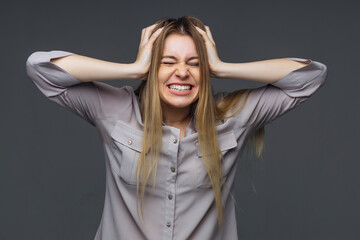 Naklejka premium Young attractive woman in a blouse on a gray background, angry, annoyed woman holds her hand behind her head. Concept of emotional people, nerves, irritation