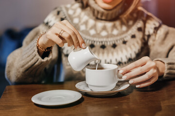 A woman in a winter sweater pours lactose-free milk or cream into a hot drink - coffee or tea. Concept of a cozy cafe and the content of animal fats in the diet