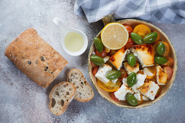 Baking form with chunks of feta cheese, tomatoes, green olives, ciabatta and olive oil over beige stone background, top view, horizontal shot