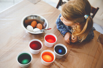 Excited little toddler girl coloring eggs for Easter. Cute happy child looking surprised at colorful colored eggs, celebrating holiday with family. From above, unrecognized face.