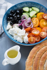 Greek salad with red and yellow tomatoes, cucumber, feta cheese, black olives and red onion served in a blue bowl, vertical shot on a grey concrete surface