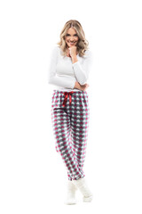 Entertained attracted young woman in pajamas watching you looking at camera with hand on chin. Full length portrait on white background.