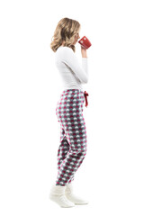 Side view of relaxed young woman in pajama and winter socks drinking coffee or tea from mug. Full length portrait on white background.