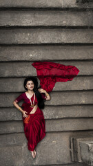 Portrait of beautiful Indian girl in heritage stepwell wearing traditional Indian red saree, gold...