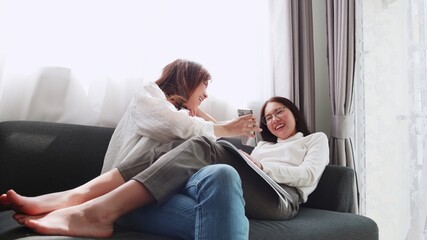 Lovely Asian couple lying on sofa play smartphone with a friend sitting and reading a book together daily holiday life of an Asian woman in the living room at home.