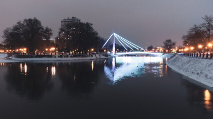 Bridge (Mist Zakokhanykh) across river in illuminated Skver Strilka in Kharkiv city center. Winter aerial evening color graded photo with water reflection