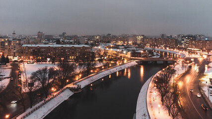 Fototapeta premium Illuminated evening riverbank with lights reflection in dark water near Skver Strilka in Kharkiv city center. Winter aerial colorful color graded photo