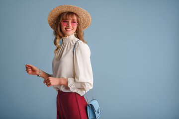 Fashion portrait of elegant happy smiling woman wearing trendy spring outfit: pink sunglasses, white blouse, straw hat, posing on blue background. Copy, empty space for text
