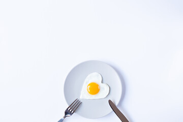 A plate with a fried egg in the form of a heart and cutlery are on a white background at the bottom of the image . Valentine's Day breakfast theme. Copy space.
