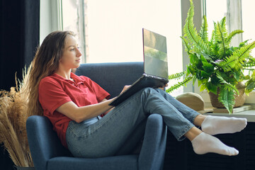 Young female using laptop after work, communicates on Internet with customer. Happy freelance woman is relaxing on comfortable couch and using laptop at home.