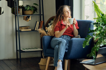 young woman with a cup of coffee in her hands sits in a chair by the window in a cozy living room