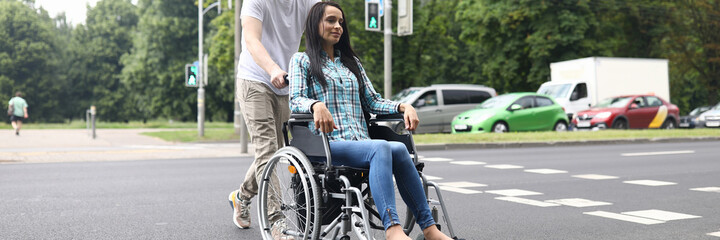 Fototapeta na wymiar A man crosses the road in the city. The guy rolls the wheelchair on which the woman is sitting