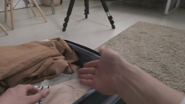 Pov of unrecognizable man sitting in living room filming video of packing suitcase, showing clothes to camera on tripod, gesturing actively