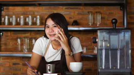 Young Asian woman barista wear apron talking and receive order from customer on cellphone at coffee shop. Concept of cafe store small business. female bartender writing note while listening client