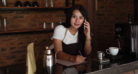 Young Asian woman barista wear apron talking and receive order from customer on cellphone at coffee shop. Concept of cafe store small business. female bartender writing note while listening client