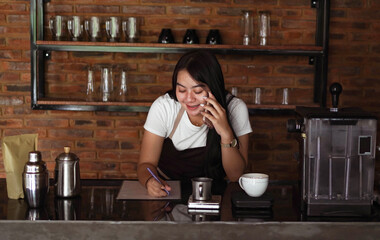 Young Asian woman barista wear apron talking and receive order from customer on cellphone at coffee shop. Concept of cafe store small business. female bartender writing note while listening client