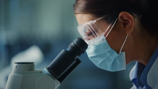 Macro Close Up Footage of a Female Scientist in Face Mask and Goggles Looking into the Microscope. Woman Microbiologist Working on Molecule Samples in Modern Laboratory with Technological Equipment.