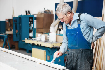 An elderly cabinetmaker in overalls and glasses paints a wooden board with a brush on a workbench...