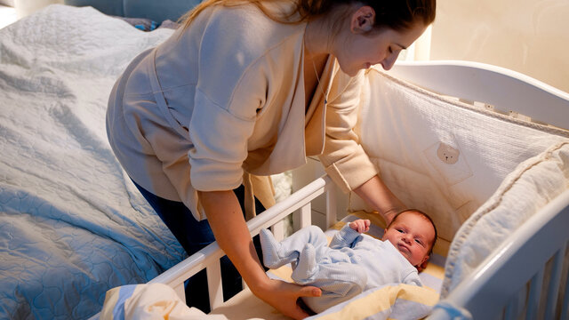 Beautiful Young Caring Mother Putting Her Newborn Baby Boy In Crib And Covering Him With Warm Blanket At Night. Concept Of Happy Parenting And Family Happiness