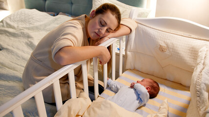 Young tired and exhausted mother fallen asleep while rocking crib of her newborn baby at night....