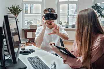Caucasian guy weared with virtual reality glasses enjoys at work with his female colleague which uses tablet in office room.