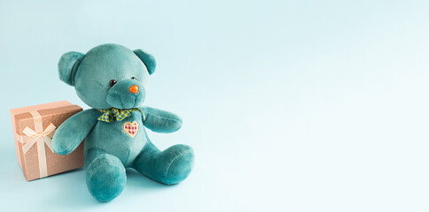 Turquoise soft teddy bear with an embroidered heart holds a gift box and a bow on a blue...