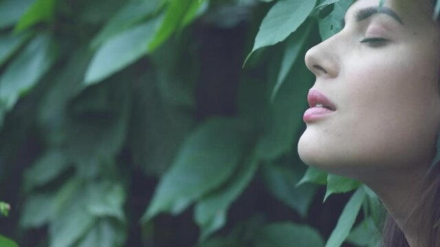 Beautiful model woman enjoying nature, breathing fresh air in summer garden over Green leaves background. Harmony concept. Enjoying nature. Slow motion 4K UHD video