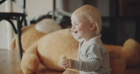 Portrait of cute baby boy smiling, eating banana, blond hair and clear blue eyes. Adorable toddler under 1 year, warm domestic background with daylight