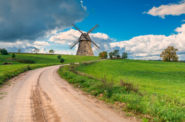 Countryside gravel road among spring fields and old windmill on horizon. Concept of ecological tourism in Baltic countries

