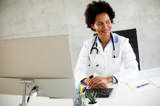 Female African American doctor wearing white coat with stethoscope sitting behind desk in office