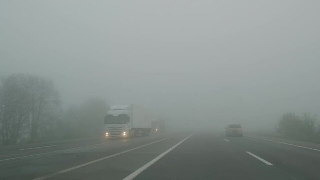 Trucks, wagons moving in fog along road, in gloomy daytime. View through frontal glass of car. Poor road visibility due to nebula. Heavy mist on highway. Concept of danger of cargo transportation. 4k
