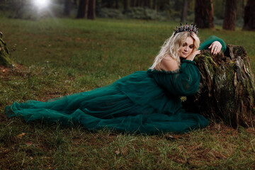 A beautiful blonde young woman in a long green dress and a diadem on her head in the forest. girl sitting near the old stump. Solar glare. Fantasy. fairy tale