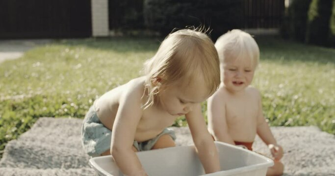Two babies are playing with bucket of foamy water at the backyard, happy childhood, Blond girl and boy on the green meadow, hot summer day out of the city. Washing carpet outdoors with a childs help.