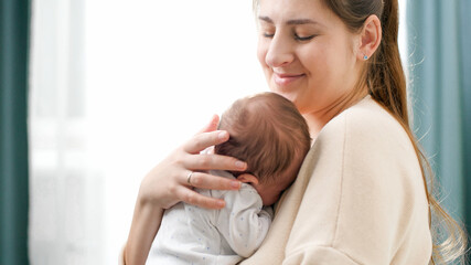 Portrait of smiling young woman cuddling and holding her newborn baby son against big window at bedroom. Concept of family happiness and loving parents with little children