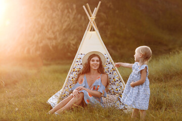 Beautiful mother with her little daughter sitting near wigwam in the field. Spending time together, outside, on vacation, outdoors. Beautiful sunset light in the garden or in the park. © Andriy Medvediuk