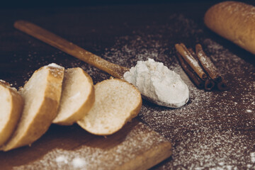 Flour in a wooden spoon, sliced French baguette on a wooden board, and cinnamon sticks sprinkled with flour on a wooden table. Side view with place for text or logo. Retro photo.