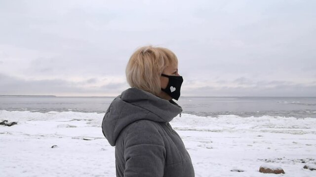 Close-up portrait of a Caucasian adult woman in a medical mask, standing against the background of the winter sea. The coronavirus epidemic. Pandemic influenza coronavirus