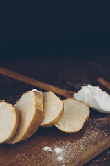 French baguette sliced on a wooden cutting board, flour in a wooden spoon dusted with flour on a wooden table, with a black background. Side view with place for text or logo. Retro photo.